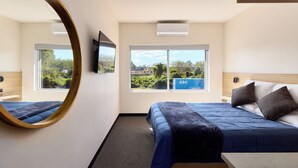 A hotel room with a large mirror, a bed with blue bedding, a television, and a window with a view of greenery and buildings. - Inala Hotel (Inala)