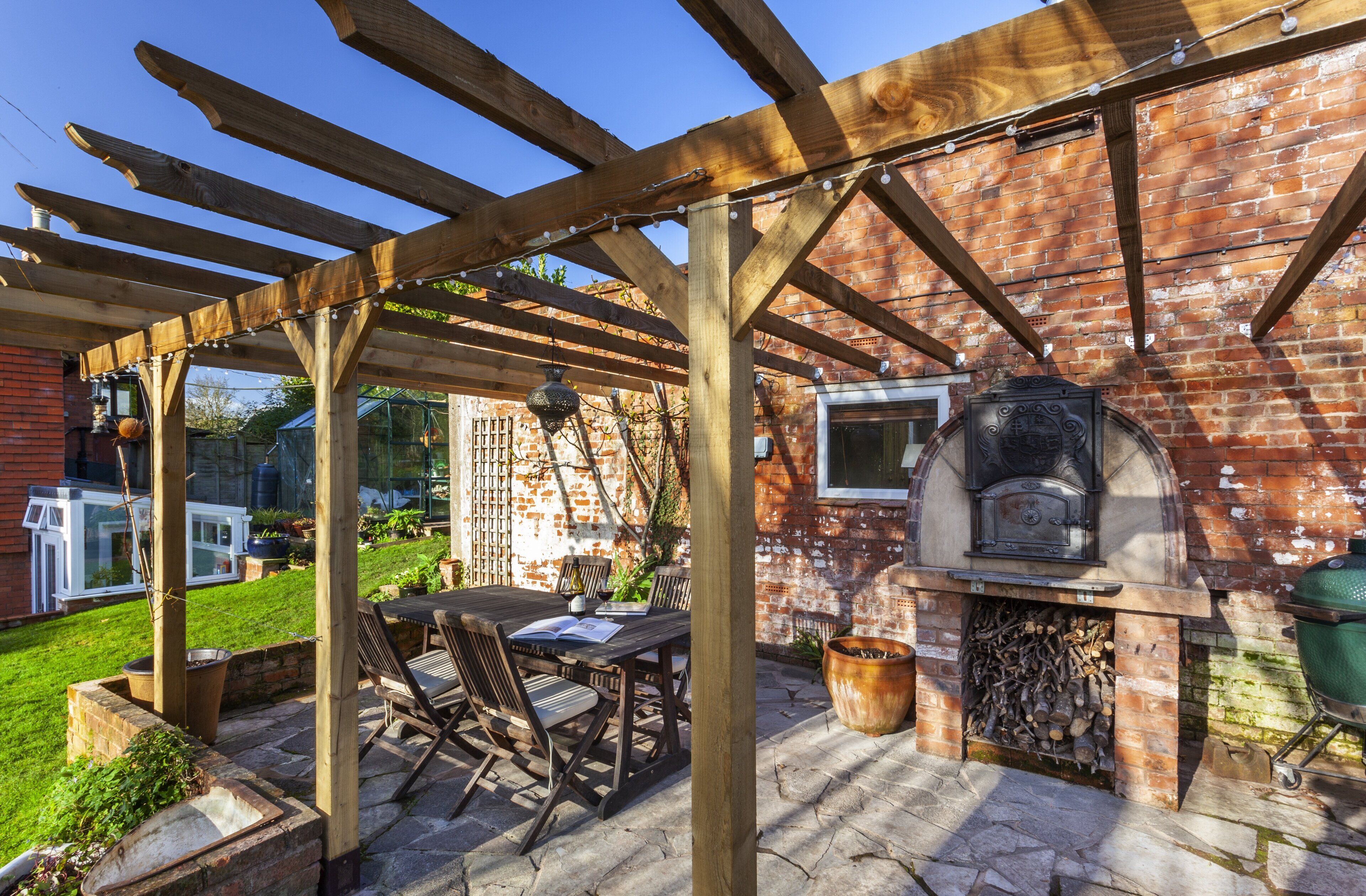 A covered patio with a brick wall, a wooden pergola, a table with chairs, and a wood-fired oven.