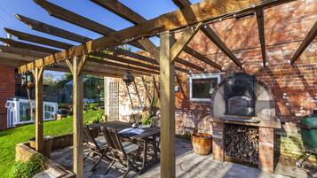 A covered patio with a brick wall, a wooden pergola, a table with chairs, and a wood-fired oven.