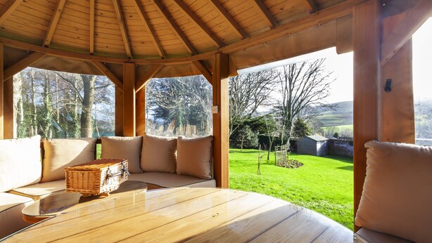 A covered outdoor seating area with a wooden table and cushions, overlooking a green landscape. - The Garden Studio, Woodcombe (Minehead)