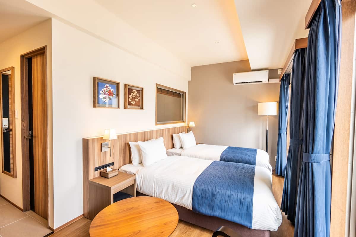 Quarto de hotel com duas camas, uma secretária de madeira, uma mesa redonda e um ar condicionado fixado na parede.