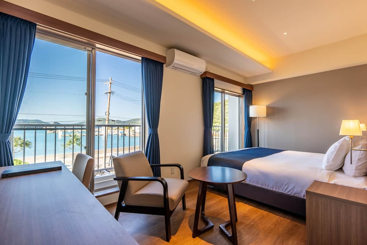 Quarto de hotel com uma grande janela, cama, escrivaninha e cadeira.