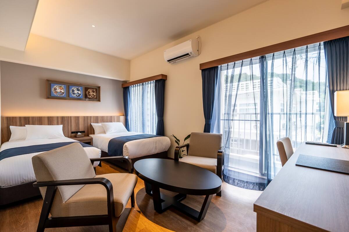 Quarto de hotel com duas camas, uma mesa de jantar, um pequeno sofá e uma escrivaninha. Há uma janela com cortinas, um aparelho de ar condicionado e quadros decorativos na parede.