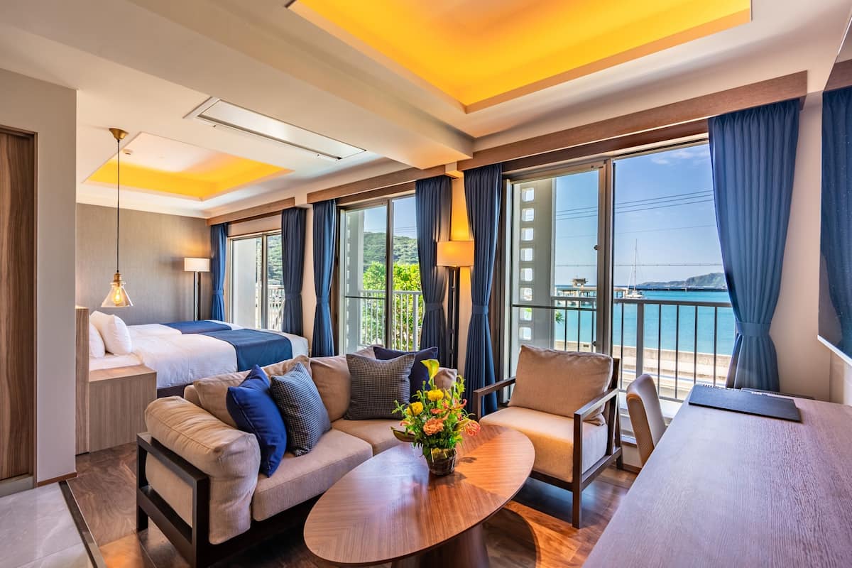 Quarto de hotel moderno com cama, sofá, poltrona e mesa de jantar. Há uma janela grande com vista para o mar e uma varanda.
