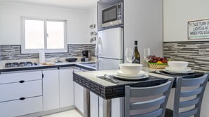 A modern kitchen with white cabinets, a built-in microwave, and a refrigerator. There is a dining table with chairs, a bowl of fruit, and a bottle of wine. - Love and Peace Vacation Rental Aruba (Noord)