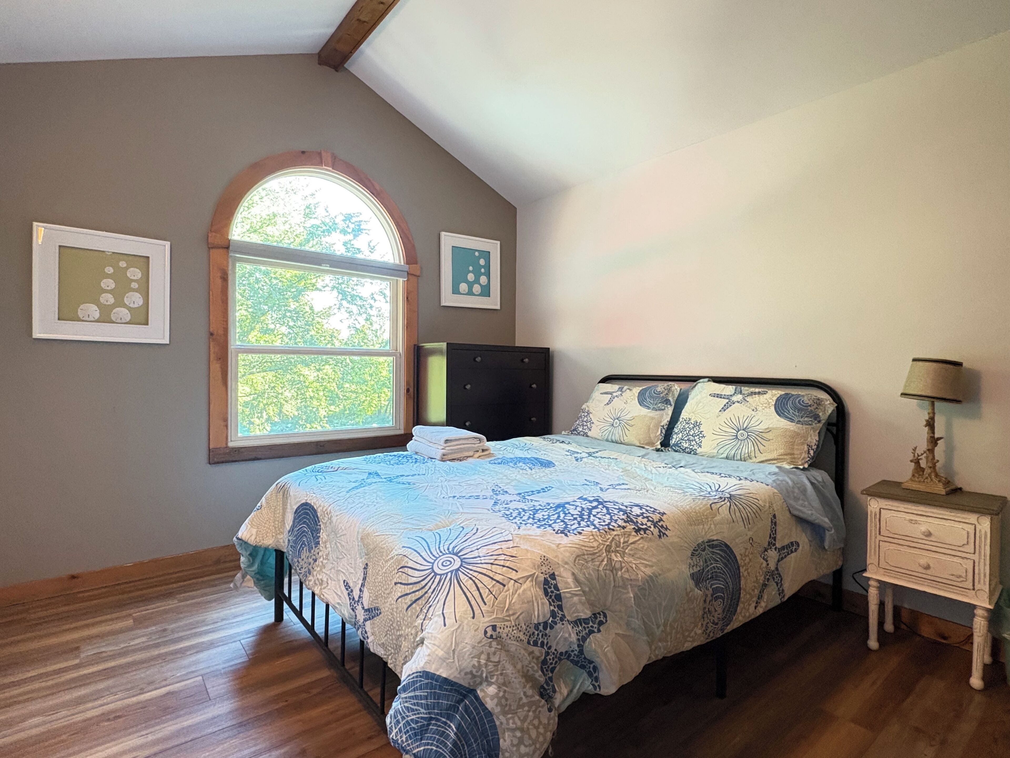 A bedroom with a bed, a nightstand, a lamp, a window, and two wall art pieces.