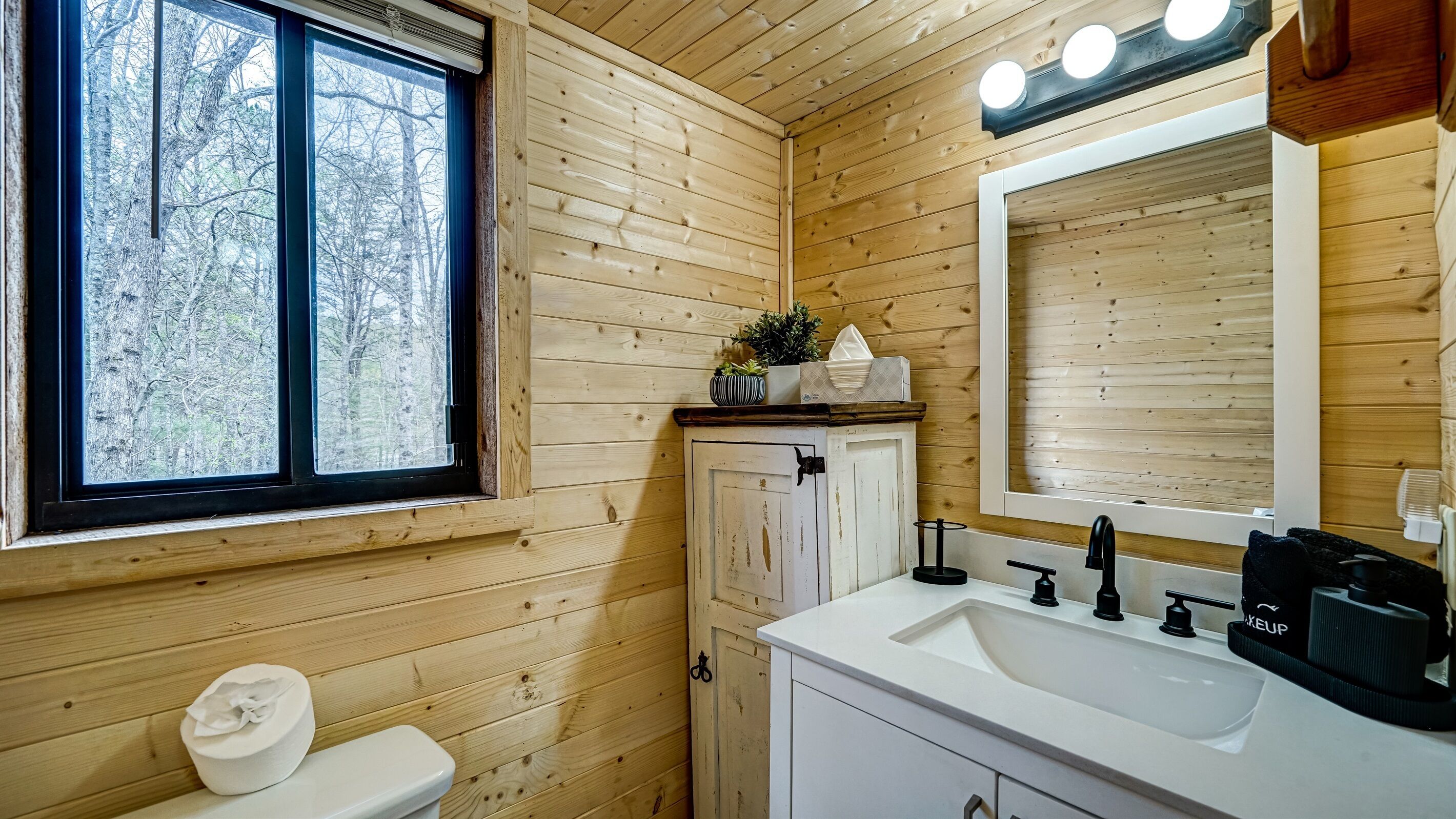 A wooden cabin bathroom with a window, a sink, a mirror, and a toilet.
