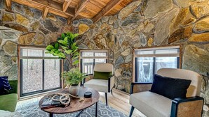 Habitación acogedora con paredes de piedra, techo de madera y dos ventanas con persianas. Hay una planta verde, una mesa redonda de madera con un libro y una guirnalda de cuentas blancas, y dos sillas.