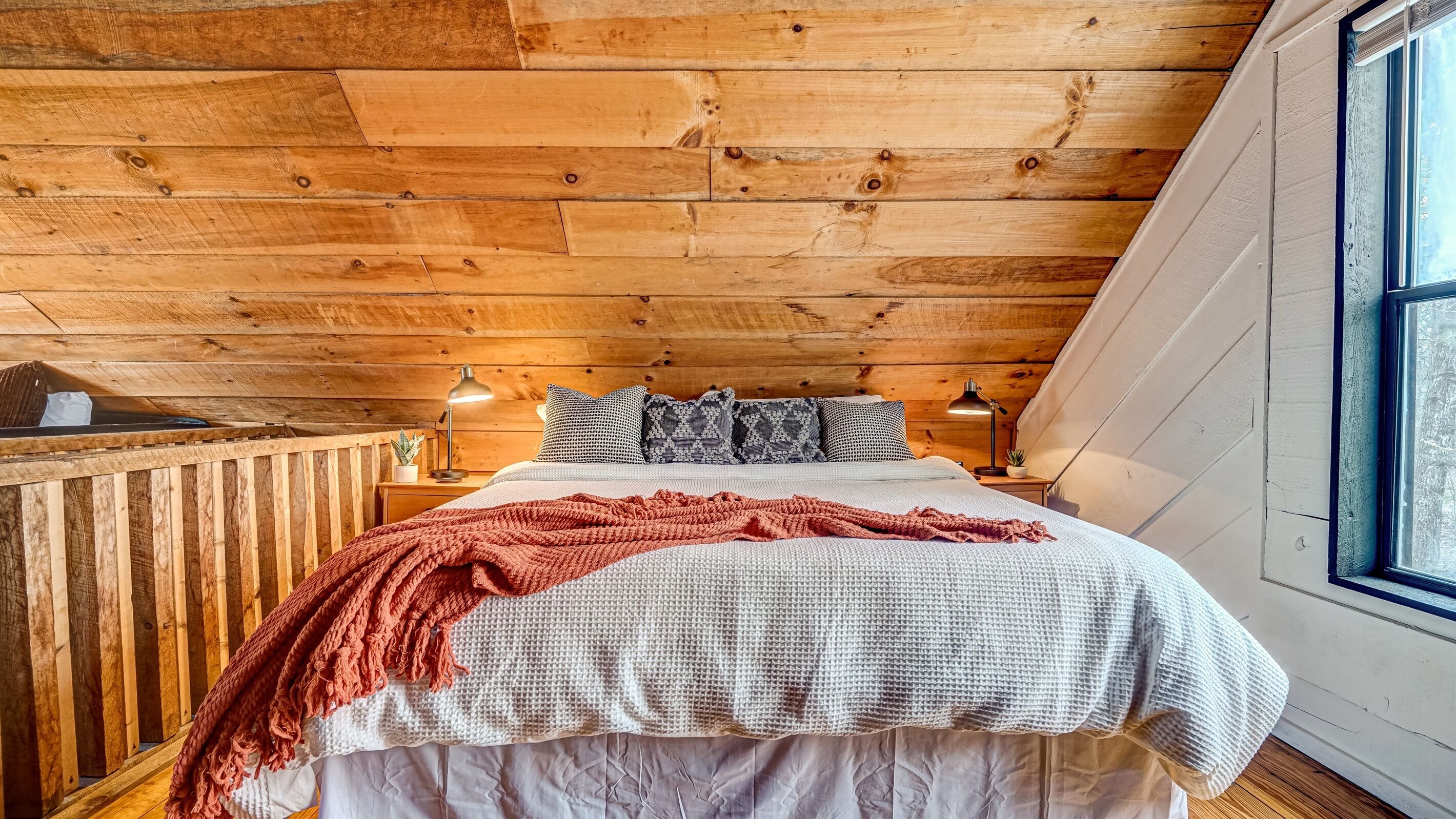 Une chambre confortable avec un plafond en bois, un lit recouvert d’une couette blanche et grise, un plaid rouge et deux lampes de chevet.