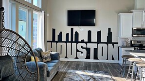 A modern living room with a large wall mural, a sofa, a chair, and a kitchen area. - Upscale brand new beautiful 3bedroom, 2 story home! (Houston)