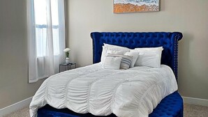 A bedroom with a large bed, a blue upholstered bench, a painting on the wall, and a window with curtains. - Upscale brand new beautiful 3bedroom, 2 story home! (Houston)