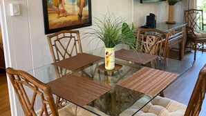 A dining area with a glass table surrounded by wicker chairs, a framed picture on the wall, and a TV mounted above the table. - Super cute, 1st floor unit across from the beach, a 1-minute walk to the beach! (Lahaina)