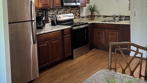A modern kitchen with wooden cabinets, stainless steel appliances, and a wooden dining table with chairs. - Super cute, 1st floor unit across from the beach, a 1-minute walk to the beach! (Lahaina)