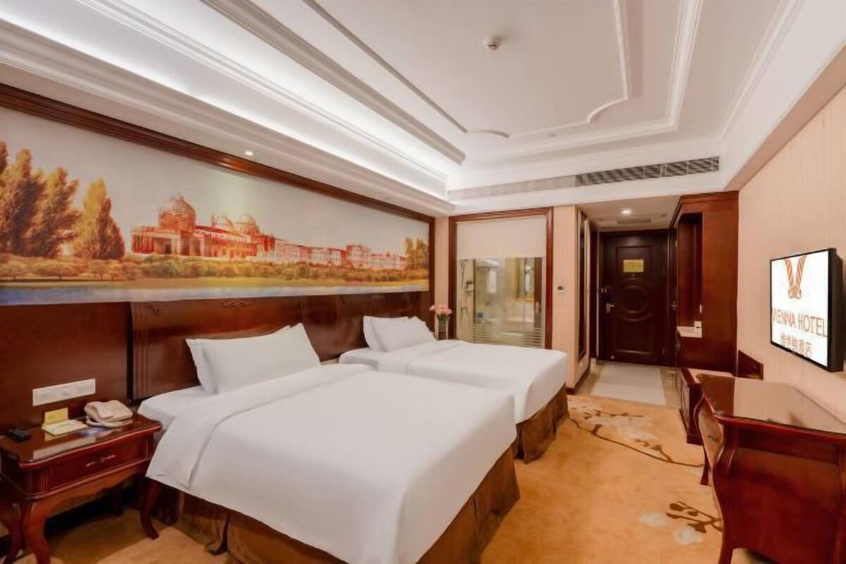 Ein Hotelzimmer mit zwei Betten, einem Schreibtisch, einem Stuhl, einem Fernseher und einem Bild an der Wand.