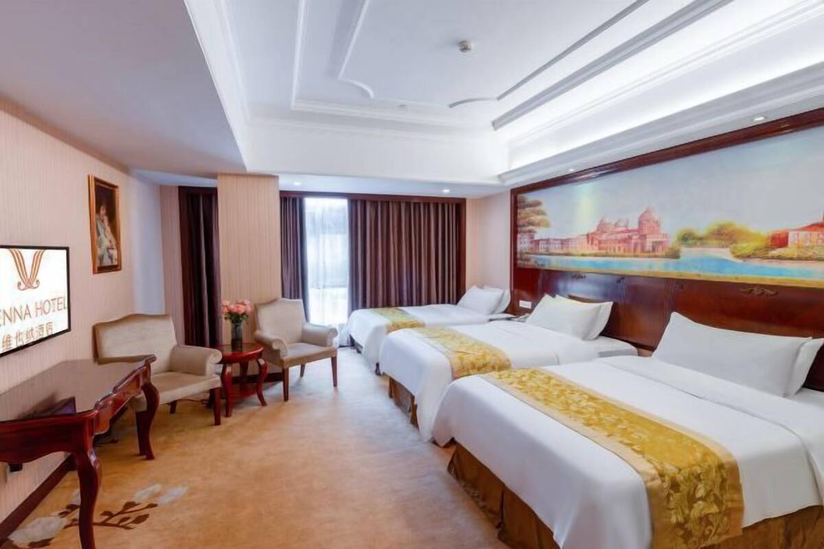 Ein Hotelzimmer mit drei Betten, einem Schreibtisch, einem Stuhl, einem Bild an der Wand und einem Fenster mit Vorhängen.
