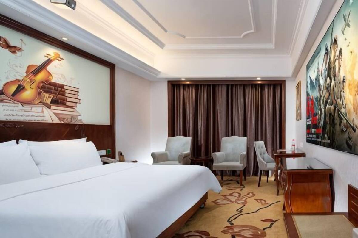 Ein Hotelzimmer mit einem großen Bett, zwei Sesseln, einem kleinen Tisch, einem Schreibtisch und einem Gemälde an der Wand.