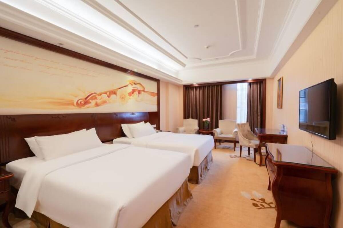 Ein Hotelzimmer mit zwei Betten, einem Fernseher, einem Schreibtisch, einem Stuhl und einem Bild an der Wand.