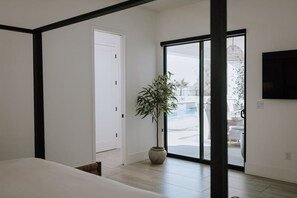 Una habitación de hotel moderna con una cama, una planta en maceta, una puerta corrediza de cristal que da a un balcón y un televisor montado en la pared.