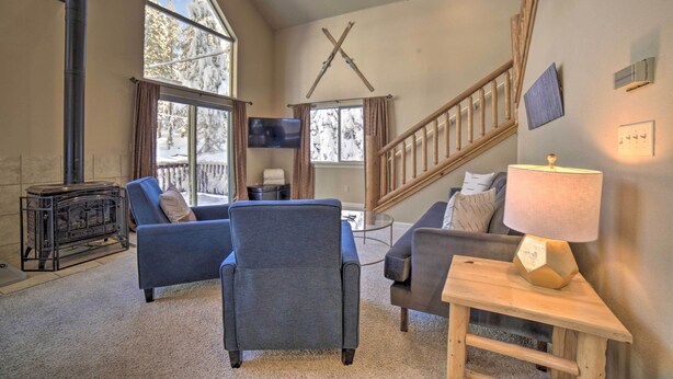 A cozy living room with a fireplace, comfortable seating, a staircase, and large windows offering a view of snow-covered trees. - Great for Groups: Soda Springs Cabin w/ Deck! (Soda Springs)