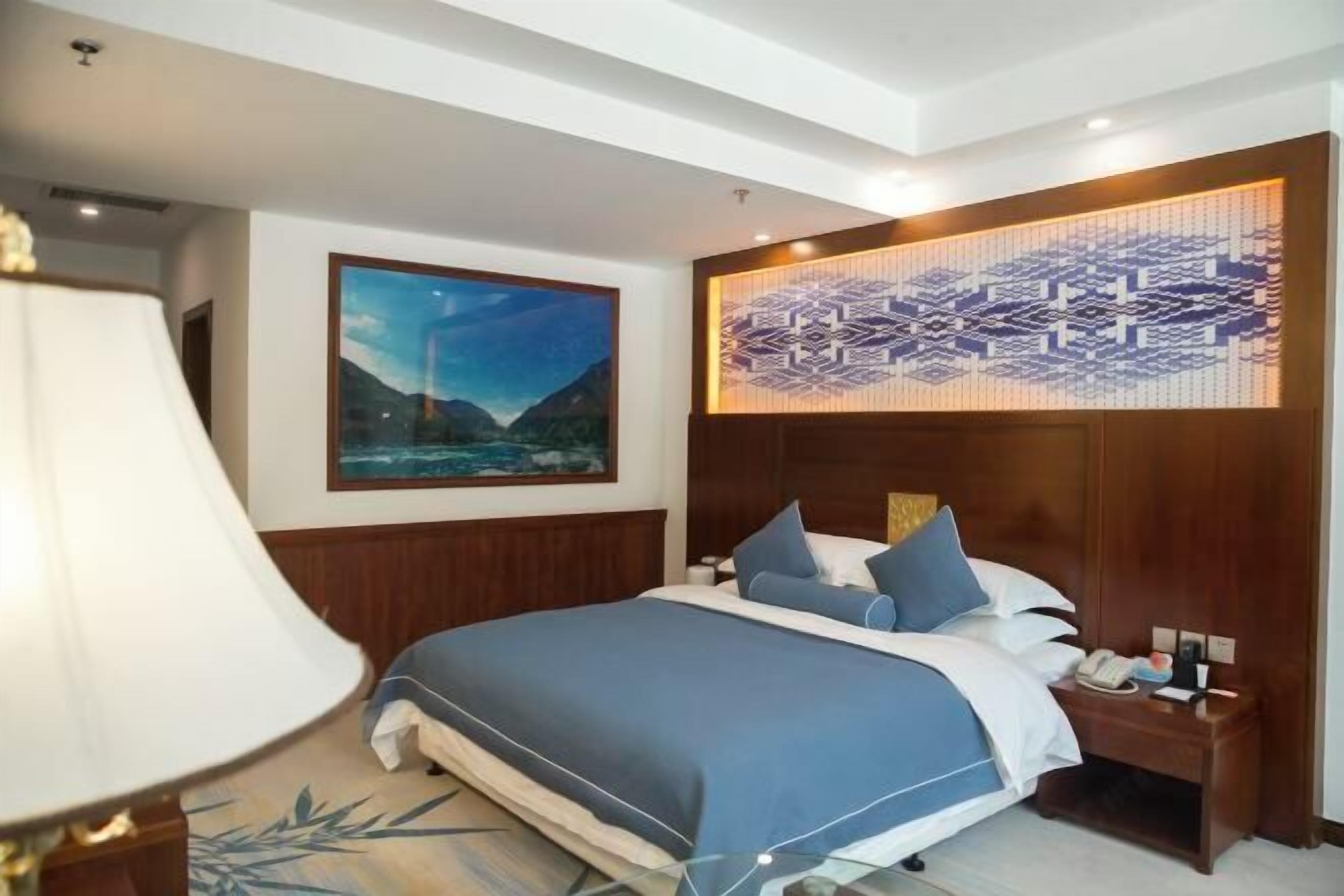A hotel room with a bed, a nightstand, a lamp, a painting, and a decorative wall panel.