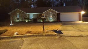 Een huis met twee verdiepingen, een gevel van natuursteen, een garage en een goed verlichte oprit in de schemering.