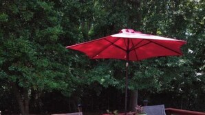 Een houten terras met een tafel, twee stoelen, een rode parasol en potplanten.