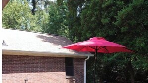 Een houten terras met een tafel en stoelen, een rode parasol, en een bakstenen gebouw op de achtergrond.
