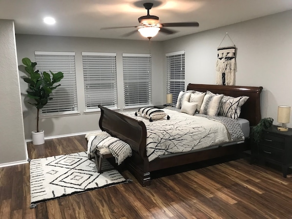 Quarto com cama, ventoinha no teto, janela com persianas, planta em vaso e tapete.