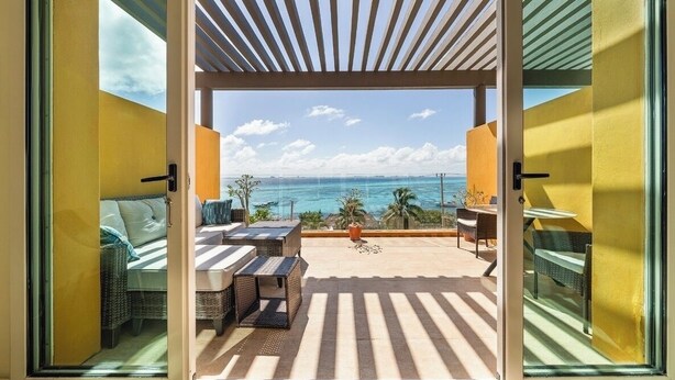 Een open deur die leidt naar een balkon met uitzicht op zee, rieten meubels en een pergola.