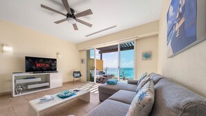 Een moderne woonkamer met een flatscreen televisie, een plafondventilator, een bank, een salontafel en uitzicht op de oceaan door grote schuifdeuren.