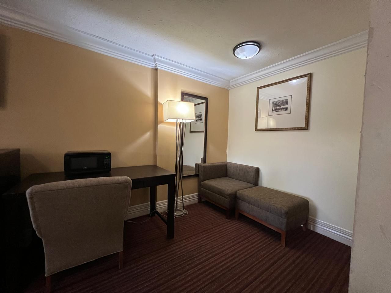 Habitación de hotel con escritorio, silla, sofá, lámpara de pie, microondas y un cuadro en la pared.