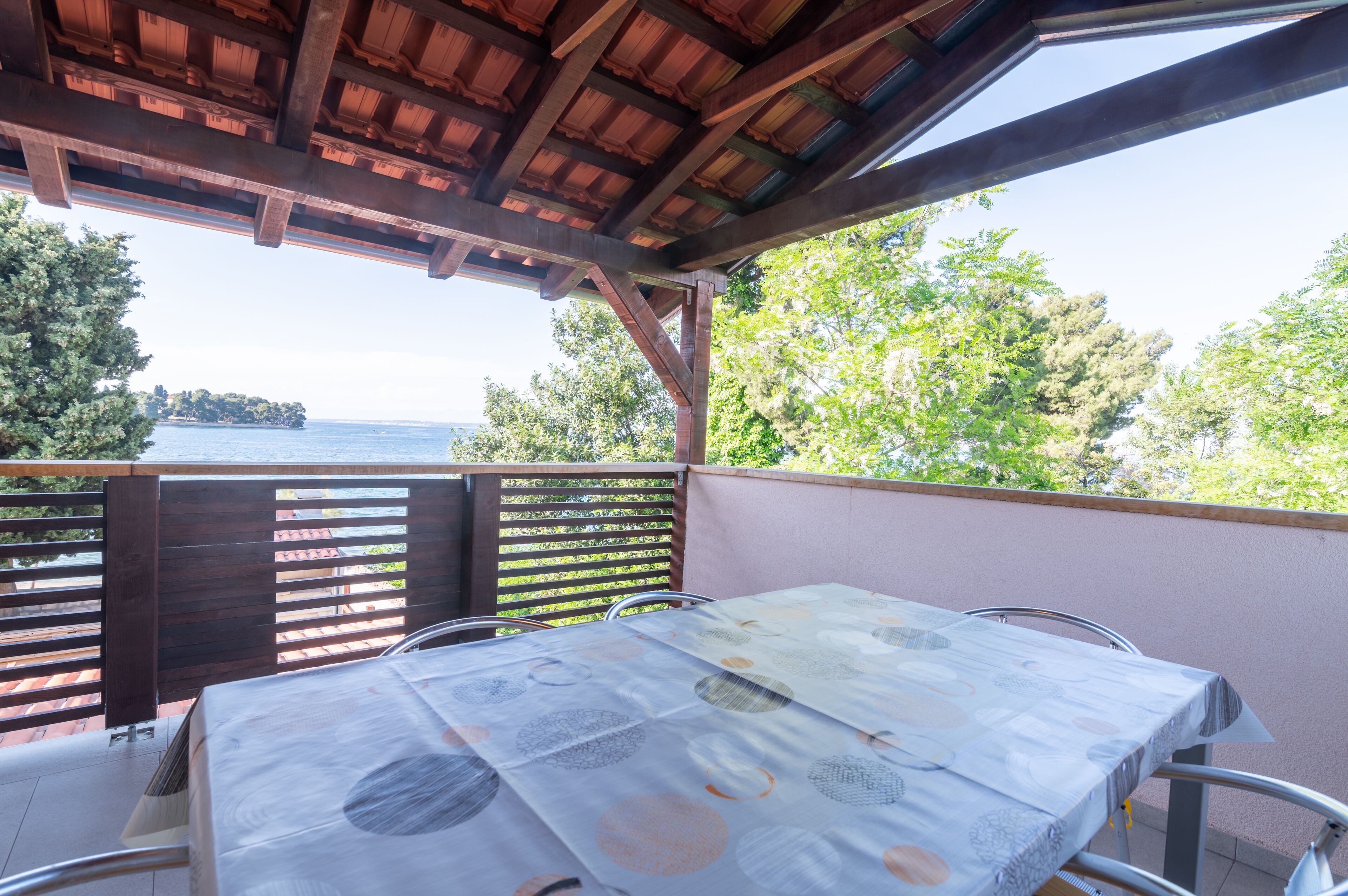 Une terrasse extérieure avec une table et des chaises, une pergola en bois et une vue sur la mer et les arbres.