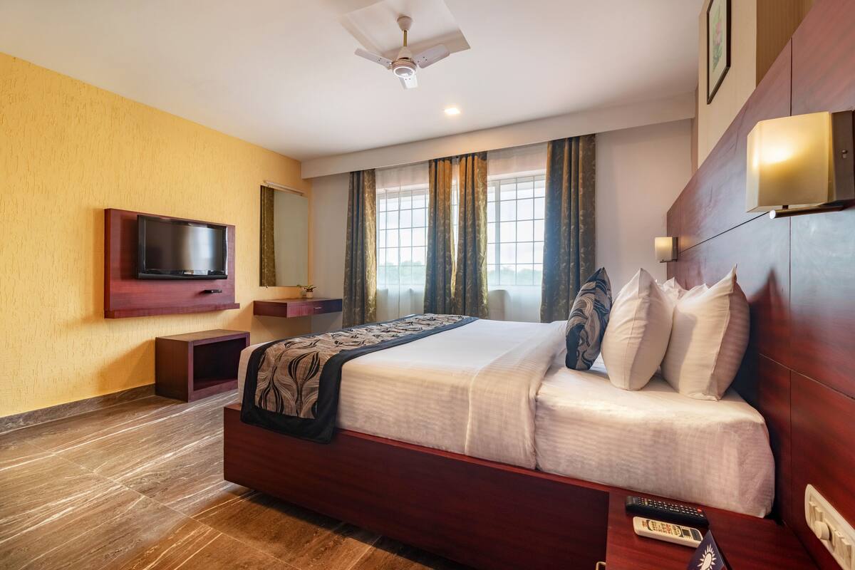 Una habitación de hotel con una cama, un televisor fijado a la pared, una mesita de noche con un control remoto y un ventilador de techo.