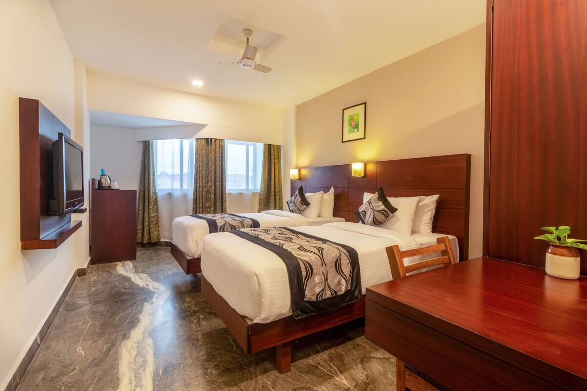 Habitación de hotel con dos camas, un escritorio, un televisor, un ventilador de techo y una ventana con cortinas.