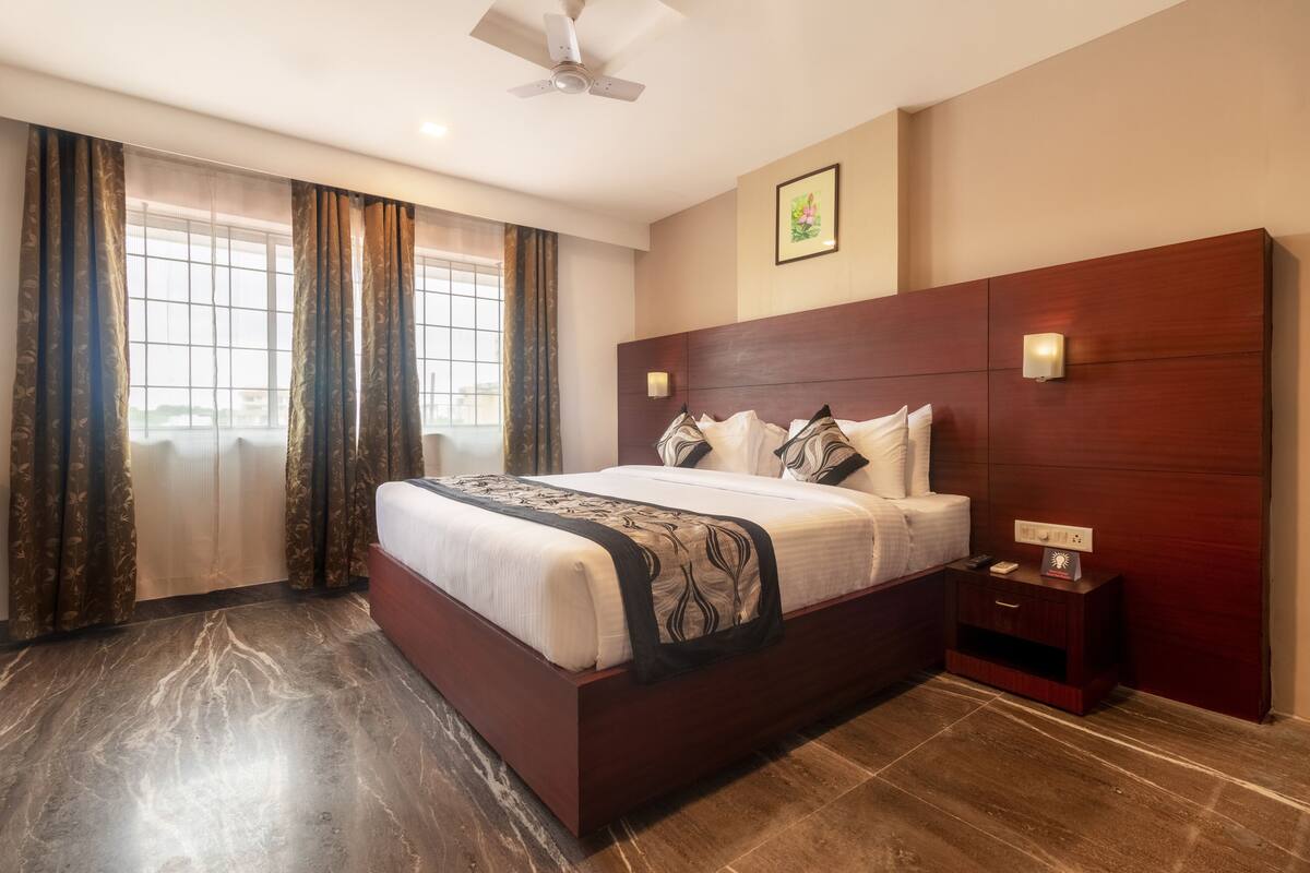 Una habitación de hotel con una cama grande, una mesita de noche, un ventilador de techo y una ventana con cortinas.