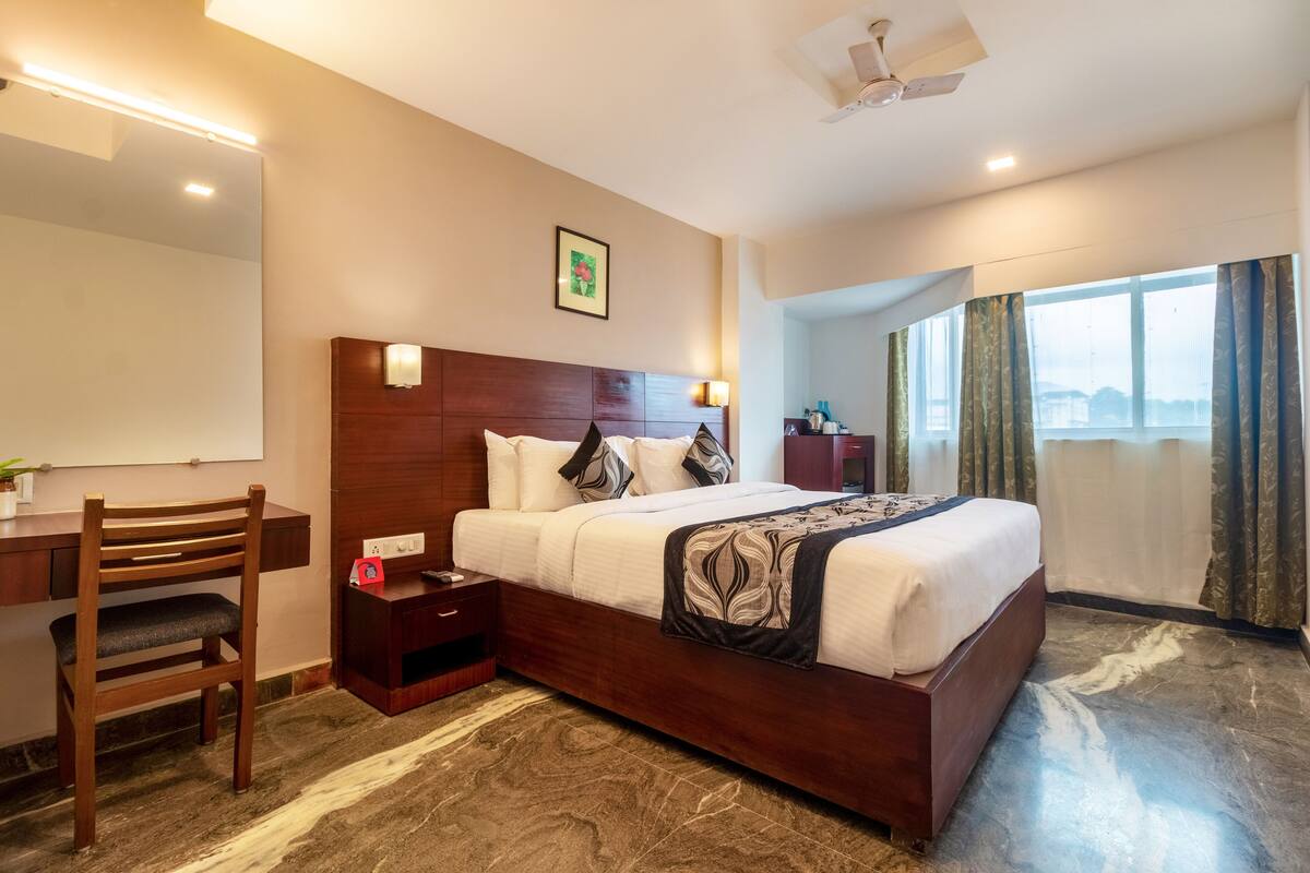 Habitación de hotel con una cama de madera, un escritorio, una silla, una mesita de noche, un espejo y una ventana con cortinas.