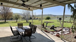 Een terras buiten met een tafel en stoelen, een pergola met een ventilator en uitzicht op een grasveld en heuvels.