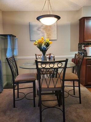 A dining area with a round glass table, four chairs, a vase with flowers, and a hanging light fixture.