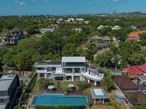 A modern two-story house with a swimming pool, surrounded by lush greenery and a hilly landscape.