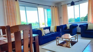 A living room with a blue sofa set, wooden chairs, a glass coffee table, and large windows with curtains. - Stay.Plus Queen's Royal Apartments Nyali (Mombasa)