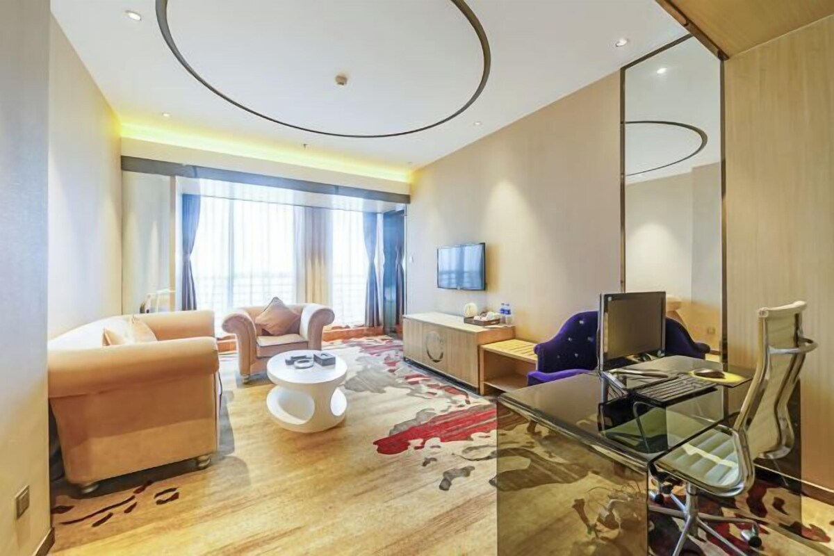 A modern hotel room with a sofa, a small round table, a glass desk with a laptop, a chair, and a large mirror.