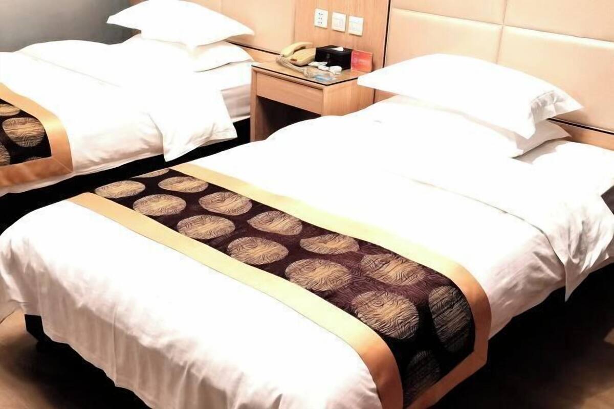 A hotel room with two beds, each with white linens and a patterned bedspread. There is a bedside table with a telephone, a cup, and a small box.