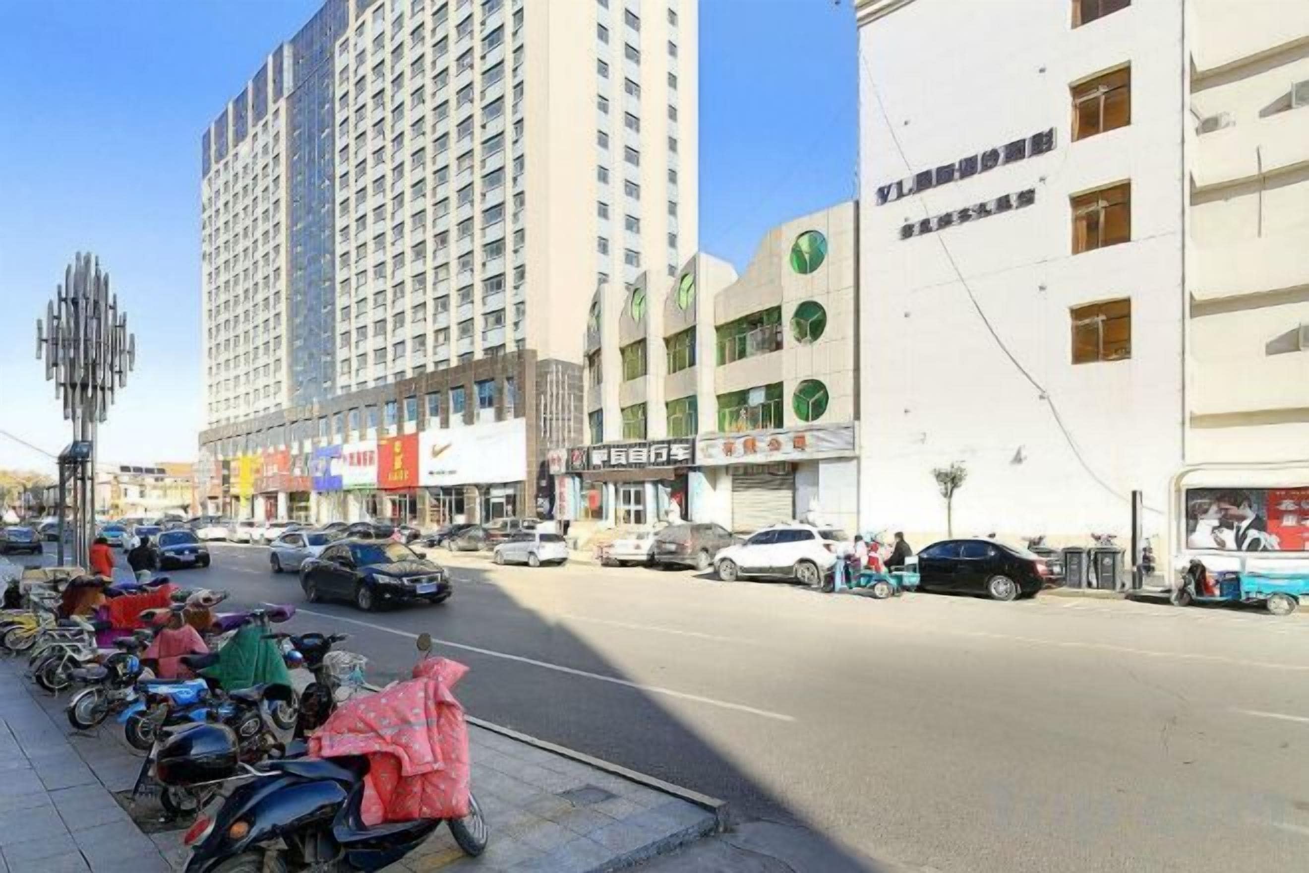 A busy urban street with tall buildings, parked scooters, and cars.