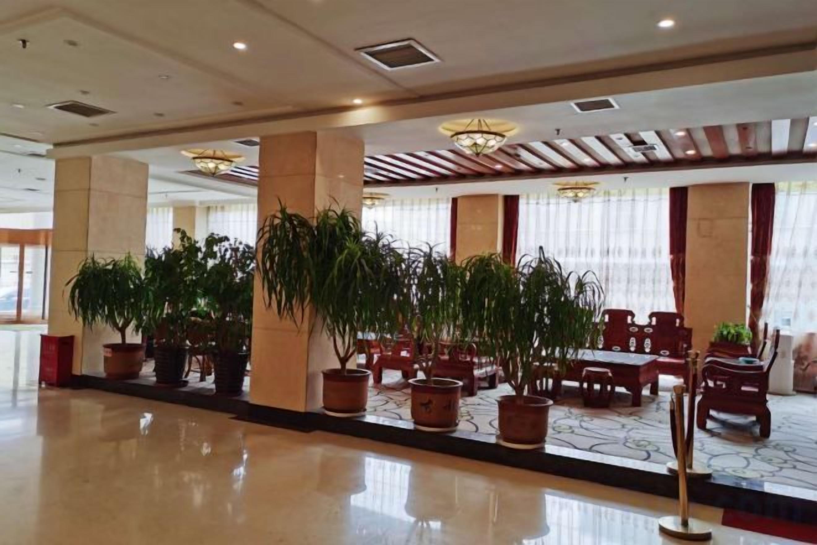 Een ruime lobby met een gepolijste vloer, grote potplanten en een zithoek met houten meubels.