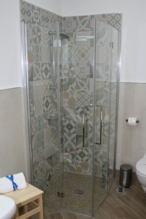 Une douche fermée par des parois en verre, avec un mur recouvert de carreaux décoratifs, un tabouret en bois et des toilettes dotées d’un porte-serviettes en papier.