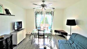 A living room with a TV, a dining table, a sofa, a ceiling fan, and a window with curtains.