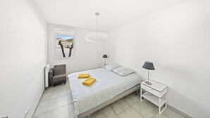 A minimalist bedroom with a bed, two bedside lamps, a chair, and a window with a view of a landscape. - Au bout de la rue la Mer T3 jardin/piscine PK/WiFi (CARQUEIRANNE)