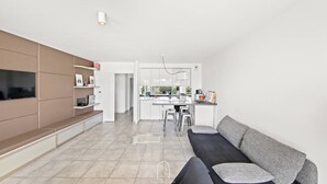 A modern living room with a sofa, a TV, and a kitchen area in the background. - Au bout de la rue la Mer T3 jardin/piscine PK/WiFi (CARQUEIRANNE)