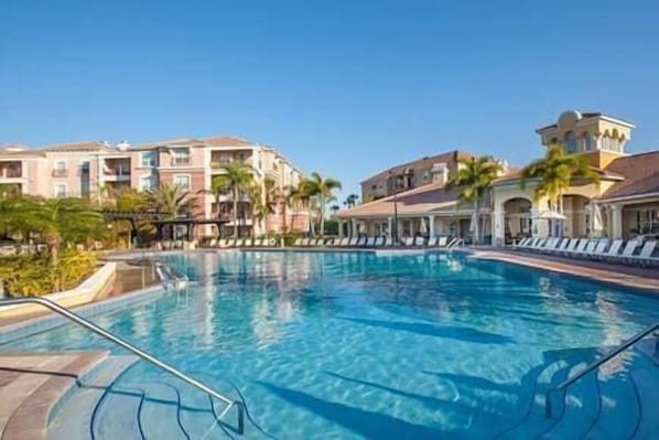 Una piscina con sillones de descanso, palmeras y edificios de varios pisos con balcones.
