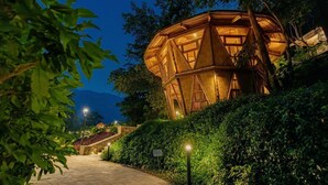 Un bâtiment en bois, de forme arrondie et coiffé d’une coupole, entouré d’une végétation luxuriante, avec un chemin pavé menant à l’entrée.